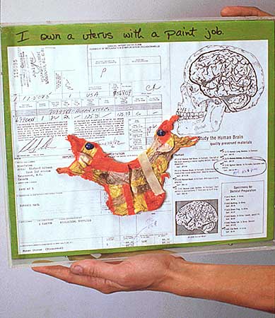 Color photo of hands holding the rectangular plaque with receipts and painted freeze-dried human uterus