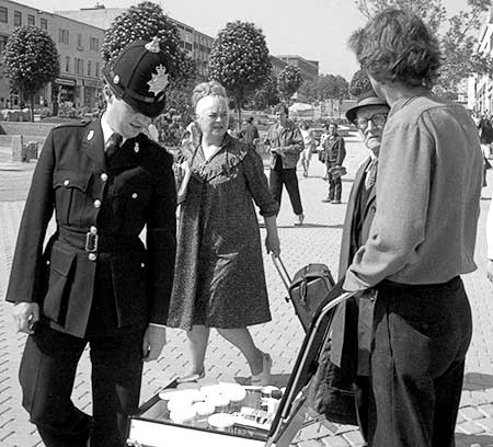 Black and white photo of a policeman looking at Gibson's display while Ms. Piddington and a pedestrian watch