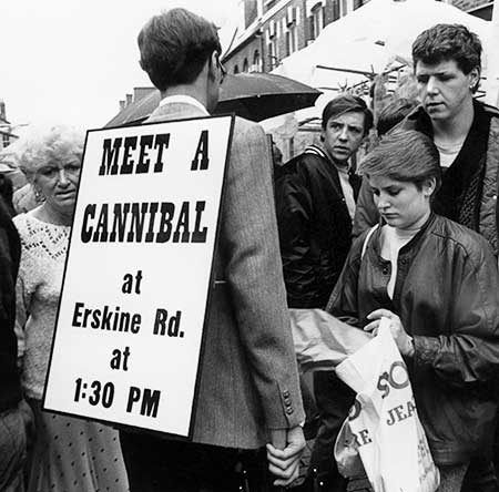 PICTURE: black and white photo of Gibson entering a crowd of shoppers wearing his sign on a rainy day.