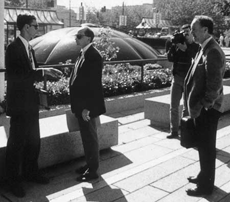 PICTURE: black and white photo of, from left to right, Gibson talking to the lead detective, a police videographer recording the event and another police detective.