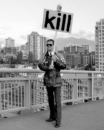 PICTURE: black and white photo of Gibson walking across the Granville Street bridge carrying a sign which reads - Kill. Photograph by Tom Buhr.